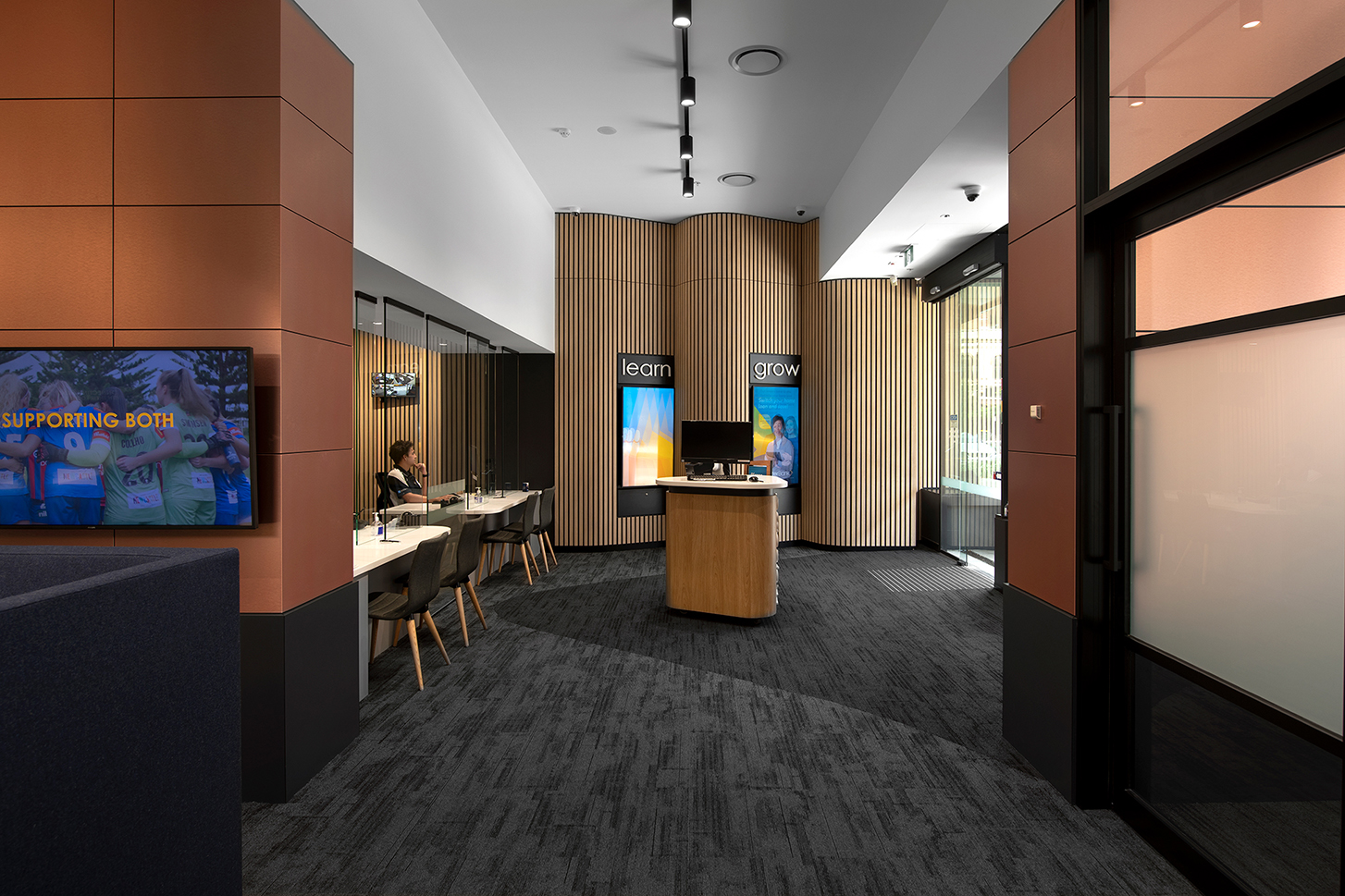 computer on island bench in middle of reception area with staff sitting behind glass panel with grey carpet and wooden accents opposite glass door entr