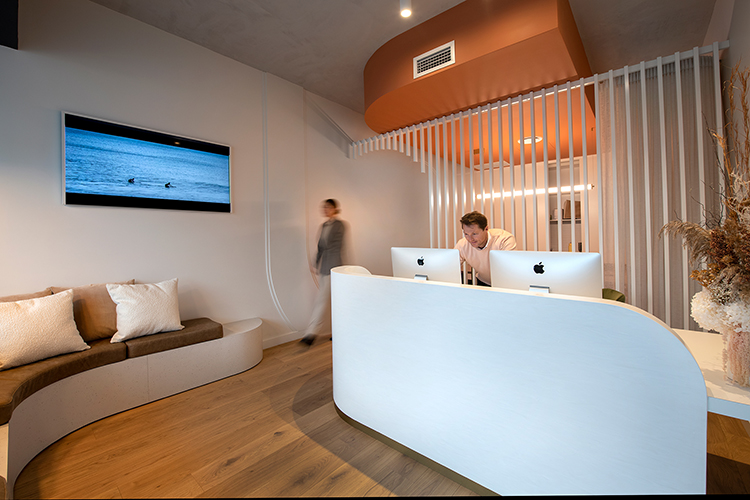 Photo of reception desk with two imacs partially visible and a staff member standing behind one operating it and a staff member in background walking from back area towards couch under tv with cushions on it